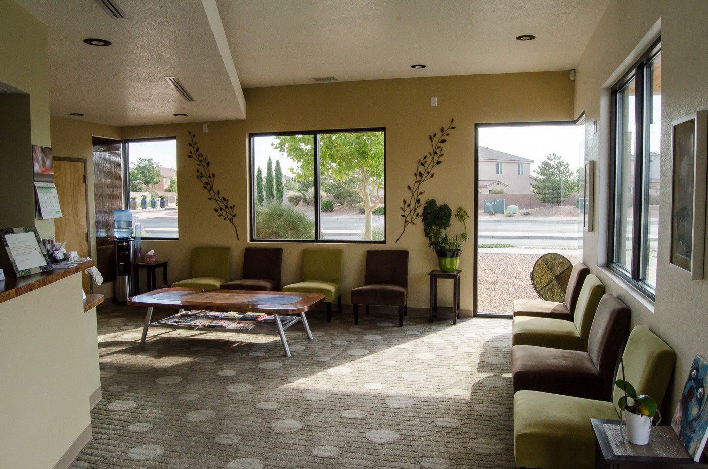 The image shows an interior space, possibly a waiting area or lobby, with modern furnishings, including seating options and a reception desk. Natural light is entering through large windows, highlighting the clean and organized environment.