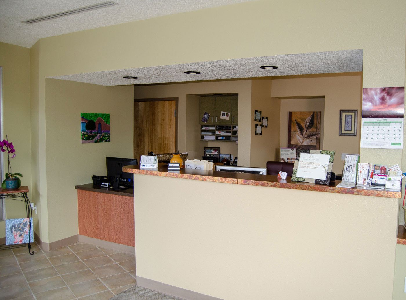 The image shows an interior view of a reception area in an office or business setting, featuring a counter with a computer monitor and keyboard, a framed picture on the wall above the counter, and a sign that reads  Welcome to our new location.