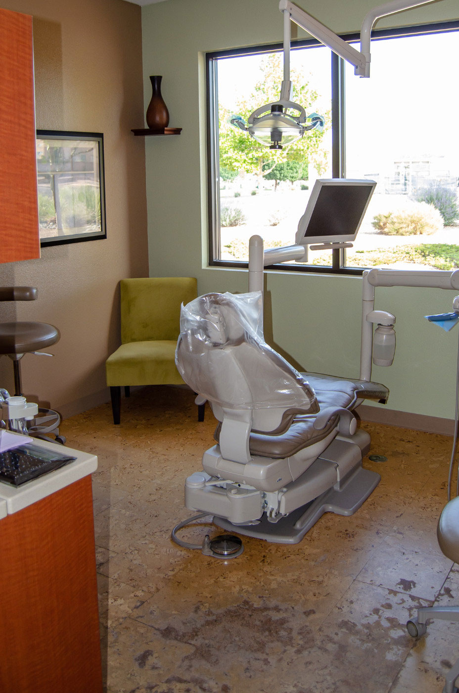 The image shows a well-equipped dental office with a chair in the foreground, a large window allowing natural light to enter, and various pieces of equipment typically found in such a setting.