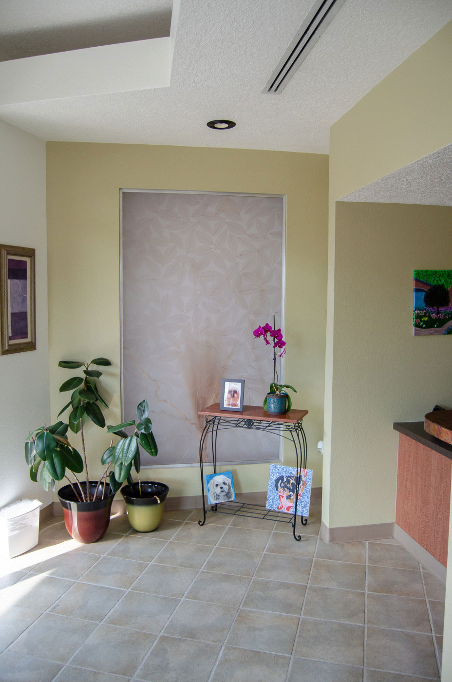 The image depicts an interior space, likely a waiting area or lobby, with a beige wall, a window covered by a blind, a plant in the corner, and a wooden floor. There is a potted plant on a table near the window, and a vase is placed on a surface to the right of the frame. The room has a light-colored carpeted floor, and there are two framed pictures hanging on the wall above the plant. The space appears clean and well-maintained.