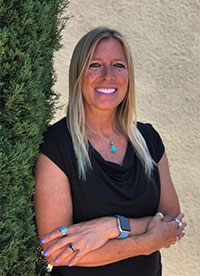 The image is a photograph of a woman standing in front of a tree, posing for the camera with her arms crossed. She has long blonde hair and is wearing a dark top.