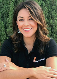 A woman in a polo shirt is smiling and leaning against a tree, wearing earrings and a ring.