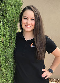 The image shows a smiling woman standing in front of a tree, posing for the camera with a friendly demeanor. She is wearing a dark top and has short hair.
