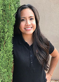 The image shows a woman standing in front of a tree, smiling at the camera.