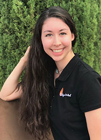 A young woman with long hair, wearing a dark polo shirt with a logo on the left chest area and a black lanyard around her neck, posing for a portrait against a background of a green hedge and a brick wall.