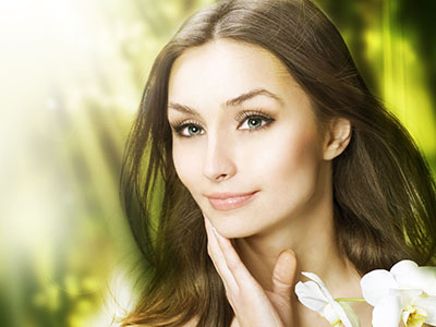 Portrait of a woman with fair skin, long dark hair, and blue eyes. She has light makeup, including lipstick, and is wearing a white top with a neckline that suggests a blouse or dress. The background features blurred greenery, giving the impression of an outdoor setting.