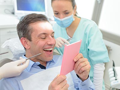 A man is seated in a dental chair, holding up a pink card with a surprised expression, while a woman in scrubs and a dental professional look on.