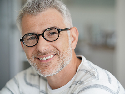 The image shows a man with glasses, smiling and looking directly at the camera. He has short hair and is wearing a white shirt with a patterned collar.