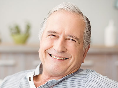 The image shows a man with white hair, wearing a blue shirt and a dark tie, smiling and looking directly at the camera. He is seated in what appears to be an indoor setting, possibly a living room or office space, as suggested by the presence of a potted plant and a wooden cabinet in the background.