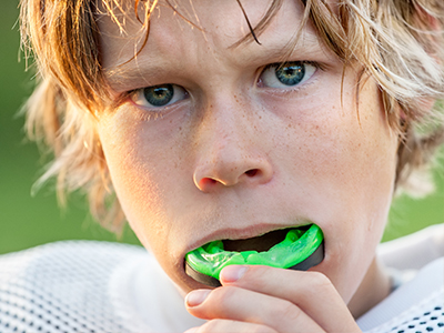 The image shows a young person with blonde hair and blue eyes, holding a green object in their mouth. They appear to be outdoors, possibly during a sports event, given the presence of what looks like a football jersey.